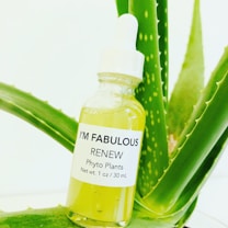 A small glass bottle with a dropper cap is filled with a yellow-green liquid. The bottle has a white label with black text that reads 'I'M FABULOUS RENEW Phyto Plants Net wt. 1 oz / 30 mL.' The bottle is positioned against the background of an aloe vera plant with green, spiky leaves.