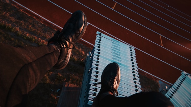 A person wearing black athletic shoes and pants is sitting on top of a stack of white sports hurdles. Below, a running track with red lanes is visible, creating a contrast with the blue and white hurdles.