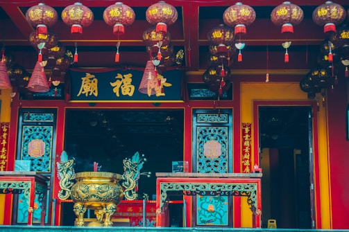 An ornate, traditional Asian temple facade adorned with vibrant red and gold colors. Numerous lanterns with Chinese characters hang from the ceiling, adding to the cultural richness. A large, intricately designed brass incense burner stands prominently in the center, flanked by decorative panels featuring ornate patterns and motifs.