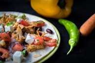 A close-up shot of a flatbread topped with roasted cauliflower, feta cheese, diced tomatoes, chopped herbs, and beans. Next to the dish, there are fresh vegetables including a yellow bell pepper, a green chili pepper, and a carrot on a dark surface.