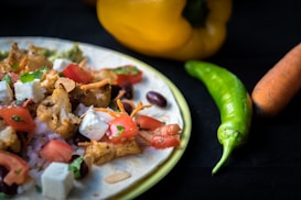 A close-up shot of a flatbread topped with roasted cauliflower, feta cheese, diced tomatoes, chopped herbs, and beans. Next to the dish, there are fresh vegetables including a yellow bell pepper, a green chili pepper, and a carrot on a dark surface.