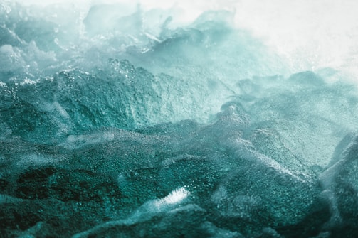 Waves of ocean water with varying shades of teal and green crashing and forming frothy textures on the surface.