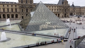 A large glass pyramid stands at the center of a courtyard surrounded by the historic Louvre Museum building. Numerous people are walking around and sitting near the pyramid, while fountains are visible in the foreground, creating an elegant water display.