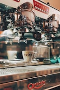 An espresso machine with chrome details and a white coffee cup with the logo 'Costa' placed on a tray. The machine has multiple buttons and a polished surface, reflecting parts of its surroundings.