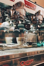 An espresso machine with chrome details and a white coffee cup with the logo 'Costa' placed on a tray. The machine has multiple buttons and a polished surface, reflecting parts of its surroundings.