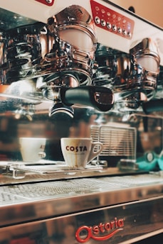 An espresso machine with chrome details and a white coffee cup with the logo 'Costa' placed on a tray. The machine has multiple buttons and a polished surface, reflecting parts of its surroundings.