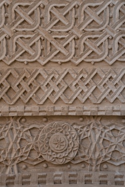 Intricate geometric patterns and floral motifs are carved into the surface of a stone wall, showcasing detailed craftsmanship. The carvings feature interlocking designs with symmetrical shapes, forming a visually complex and harmonious texture.