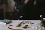 A wooden incense holder with a thin, burning incense stick placed on a table. Wisps of smoke are rising delicately from the incense. There are small piles of dried herbs and possibly seeds scattered around. A book lies closed on the table in the background, and the overall scene is softly lit with a dark, blurred background.