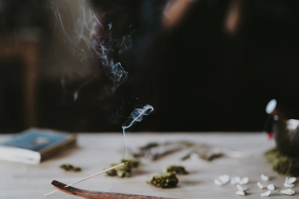 A wooden incense holder with a thin, burning incense stick placed on a table. Wisps of smoke are rising delicately from the incense. There are small piles of dried herbs and possibly seeds scattered around. A book lies closed on the table in the background, and the overall scene is softly lit with a dark, blurred background.