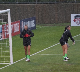 Two soccer players, dressed in black training kits with green socks and cleats, are standing on a wet soccer field near a goalpost. It is raining, and one player appears to be smiling and clapping, while the other is stretching or warming up. The background includes advertisements and a sign bearing a soccer logo.