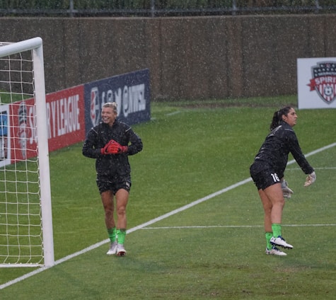 Two soccer players, dressed in black training kits with green socks and cleats, are standing on a wet soccer field near a goalpost. It is raining, and one player appears to be smiling and clapping, while the other is stretching or warming up. The background includes advertisements and a sign bearing a soccer logo.