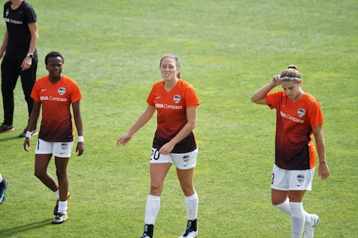 Three female soccer players walk across a grassy field wearing orange and black team jerseys with the logo 'DASH' and sponsor 'BBVA Compass'. They appear to be focused, possibly after a training session or match.