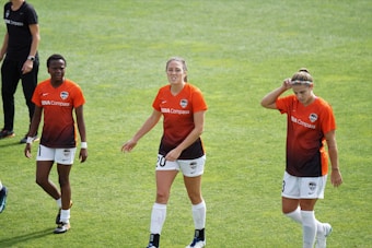 Three female soccer players walk across a grassy field wearing orange and black team jerseys with the logo 'DASH' and sponsor 'BBVA Compass'. They appear to be focused, possibly after a training session or match.