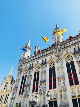 A historic Gothic-style building with ornate stone carvings and arched windows adorned with red frames. Vibrant flags are flying atop the spires against a clear blue sky, and the facade includes intricate decorative crests and statues.