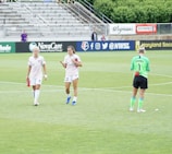 Three athletes are on a soccer field; two are wearing white sports uniforms while one is in a green goalkeeper outfit. They appear to be walking across the field, with a background of empty stands and various advertisements.