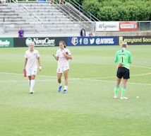 Three athletes are on a soccer field; two are wearing white sports uniforms while one is in a green goalkeeper outfit. They appear to be walking across the field, with a background of empty stands and various advertisements.
