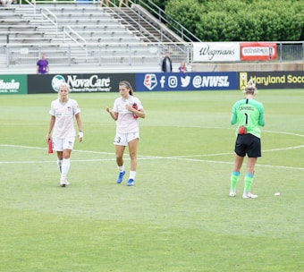 Three athletes are on a soccer field; two are wearing white sports uniforms while one is in a green goalkeeper outfit. They appear to be walking across the field, with a background of empty stands and various advertisements.