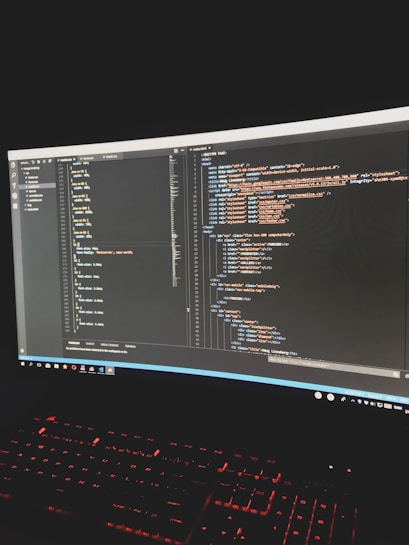 A dark room illuminated by the glow of a computer monitor displaying HTML code. The screen shows a text editor with two open documents containing structured code. Below the monitor is a keyboard with illuminated red keys.