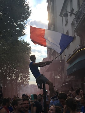 A person is balanced on a pole, holding a large French flag. The scene is a lively street with a crowd of cheering people. Sunlight filters through the clouds and trees, creating a dynamic backdrop.