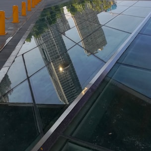 A glass surface reflects the image of tall buildings and green trees, creating an illusion of the structures and foliage being upside down. There are yellow bollards aligned on a paved path to the left of the glass.