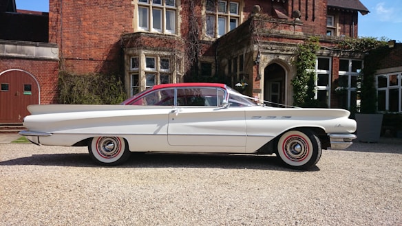 A classic white car with a red roof is parked in front of an ornate brick building. The car has distinctive tail fins and whitewall tires, reflecting vintage design. The building features tall windows, ivy on the walls, and an arched doorway, suggesting historical or architectural significance.