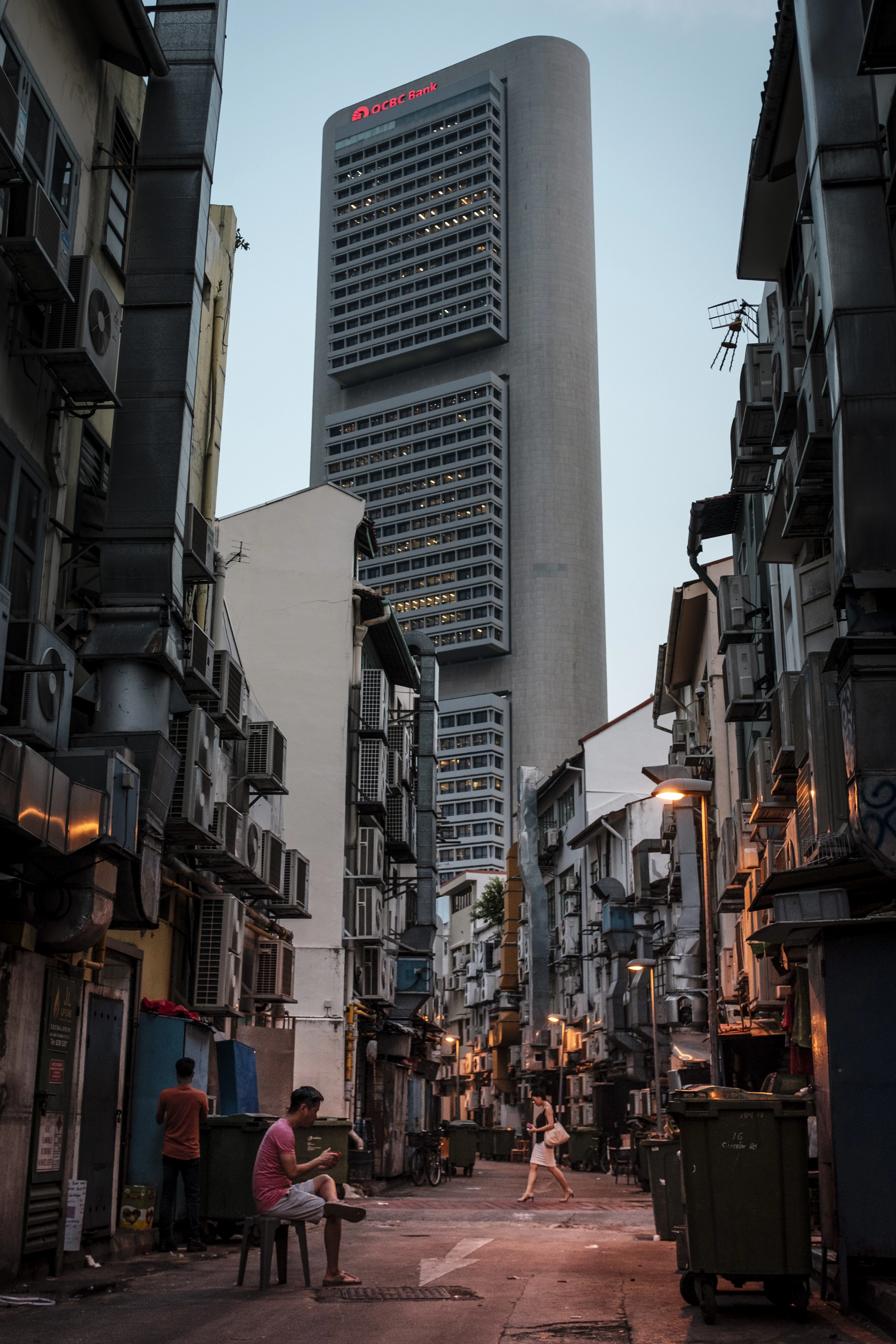 A narrow Chinatown backstreet lined with shophouses and air-con units, a skyscraper towering behind at dusk.