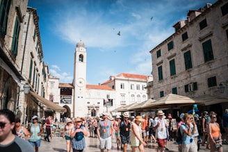A bustling historic square filled with tourists, surrounded by old stone buildings and a prominent clock tower. People are wearing summer clothing, and some are taking photographs. The sky is bright blue with a few clouds and birds flying above.