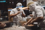 Two chefs wearing white uniforms and masks are focused on preparing dough and filling for dumplings in a dimly lit kitchen. In the foreground, one chef is handling a dough sheet, while the other is working with filling. Wooden steamers are stacked on the counter, suggesting a busy and traditional kitchen setup.