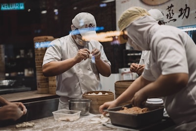 Two chefs wearing white uniforms and masks are focused on preparing dough and filling for dumplings in a dimly lit kitchen. In the foreground, one chef is handling a dough sheet, while the other is working with filling. Wooden steamers are stacked on the counter, suggesting a busy and traditional kitchen setup.