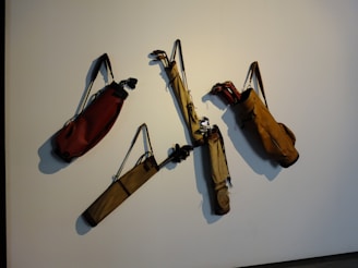 Several golf bags with straps are hanging on a wall, each containing a few golf clubs. The bags are arranged in a scattered manner on a plain background.