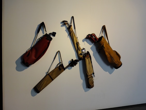 Several golf bags with straps are hanging on a wall, each containing a few golf clubs. The bags are arranged in a scattered manner on a plain background.