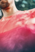 A person wearing a brightly colored shirt with a gradient pattern, transitioning from coral to plum. The shirt has a small, visible brand logo. The background is blurred with green tones, suggestive of foliage.