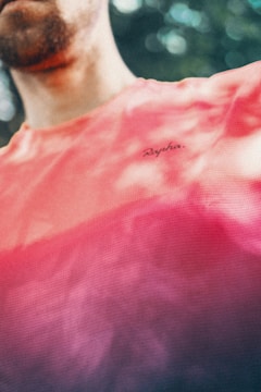 A person wearing a brightly colored shirt with a gradient pattern, transitioning from coral to plum. The shirt has a small, visible brand logo. The background is blurred with green tones, suggestive of foliage.