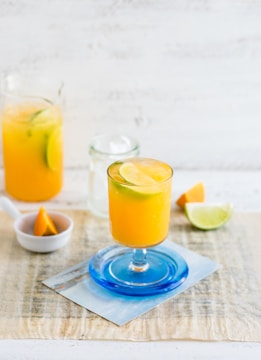 A vibrant orange drink garnished with lime slices is served in a glass with a blue base. Nearby, there's a clear jug filled with a similar orange beverage and lime wedges. A small dish holds an orange wedge, and slices of lime and orange rest on the light-colored surface.