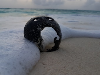 A weathered, dark metallic object partially submerged in the sand is surrounded by white sea foam as ocean waves gently wash over it. The sea and cloudy sky form a serene and moody background, adding to the mystique.