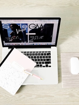 A laptop displaying a webpage with various tabs open. In front of the laptop, there is a closed notebook with a speckled cover and a pink pen resting on it. A white wireless mouse is positioned on the right side of the laptop, all set on a light wooden surface.