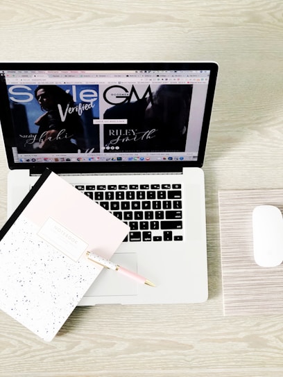 A laptop displaying a webpage with various tabs open. In front of the laptop, there is a closed notebook with a speckled cover and a pink pen resting on it. A white wireless mouse is positioned on the right side of the laptop, all set on a light wooden surface.