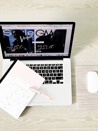 A laptop displaying a webpage with various tabs open. In front of the laptop, there is a closed notebook with a speckled cover and a pink pen resting on it. A white wireless mouse is positioned on the right side of the laptop, all set on a light wooden surface.