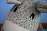 A close-up of a plush toy featuring a textured, furry surface. The toy has large, shiny black eyes and floppy ears. The background is a plain light blue wall.