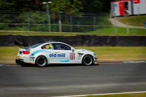 A race car adorned with various sponsor logos is shown speeding along a racetrack. The vehicle is predominantly white with blue and red accents, featuring prominent logos on the side. In the background, there is a grassy area bordered by a wire fence, and some trees are visible.