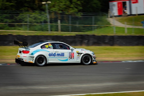 A race car adorned with various sponsor logos is shown speeding along a racetrack. The vehicle is predominantly white with blue and red accents, featuring prominent logos on the side. In the background, there is a grassy area bordered by a wire fence, and some trees are visible.