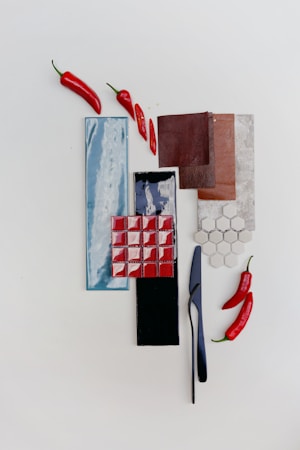 The image consists of an arrangement of colorful ceramic tiles, leather swatches, and red chili peppers placed on a white surface. The tiles include a long blue rectangular one, a black one, small red square tiles, and small white hexagonal tiles. Three pieces of brown leather or fabric are stacked together. A black knife is also part of the arrangement.