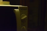 A close-up view of a microwave oven's control panel with two rotary dials and a brand name 'Techwood'. The lighting is dim, creating a shadowy ambiance.