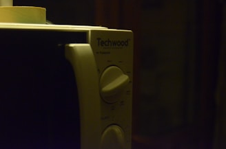 A close-up view of a microwave oven's control panel with two rotary dials and a brand name 'Techwood'. The lighting is dim, creating a shadowy ambiance.