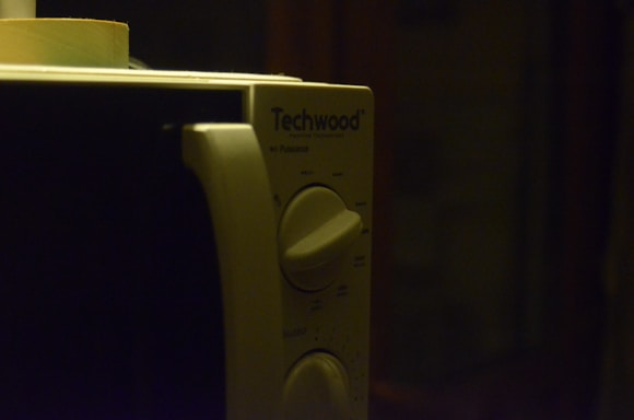 A close-up view of a microwave oven's control panel with two rotary dials and a brand name 'Techwood'. The lighting is dim, creating a shadowy ambiance.