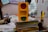 A handmade yellow traffic light model with red, yellow, and green circles is placed on a stack of books and papers. The background includes people engaged in activities, educational posters, and various classroom materials.