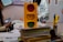 A handmade yellow traffic light model with red, yellow, and green circles is placed on a stack of books and papers. The background includes people engaged in activities, educational posters, and various classroom materials.