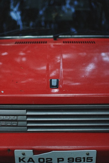 A vintage red car hood with a badge labeled 'RUTI 800' on a chrome grille. The car's license plate shows 'KA 02 P 9615'. The image features reflections of trees above, creating an outdoor setting.