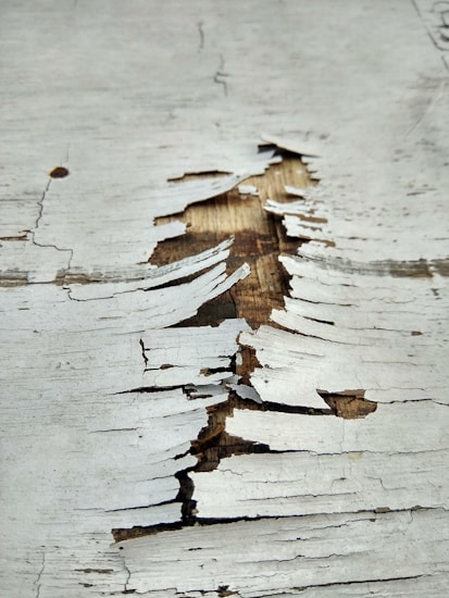 Peeling white paint on a wooden surface reveals the brown wood underneath. The paint is cracked and flaked in various sizes, creating a textured appearance.