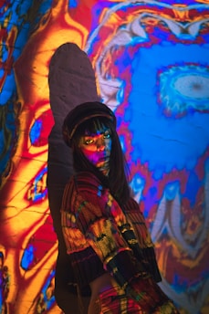 A person stands against a wall covered with psychedelic, colorful projections. The individual is wearing a multicolored sweater and a hat, with vibrant colors such as red, blue, and yellow cast onto them. Shadows and light interact to create an abstract and vivid atmosphere.